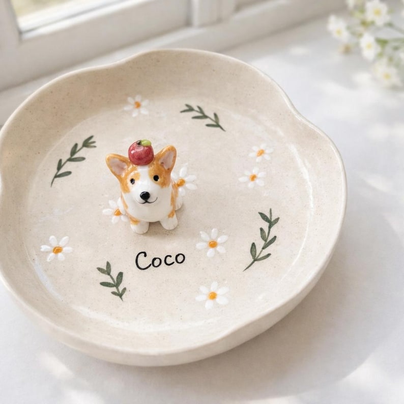 May include: A beige ceramic dish with a raised rim, featuring a small corgi figurine wearing a red apple on its head. The dish is decorated with white daisies and green sprigs, with the name "Coco" written in black.