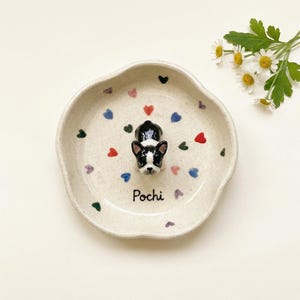 May include: A ceramic dish featuring a Boston Terrier figurine. The off-white dish has a wavy edge and is decorated with colourful hearts. The word "Pochi" is written on the dish.