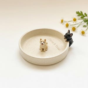 May include: A beige ceramic dish with two dog figurines. One is a light brown Shiba Inu, and the other is a black French Bulldog. The dish has the words "Hana & Yuzu" inscribed on it. Daisies are in the background.