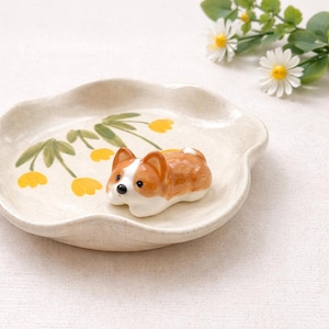 Puede incluir: Una figura de corgi de cerámica con pelaje marrón y blanco descansa sobre un plato crema con volantes. El plato está decorado con flores amarillas y hojas verdes. El corgi tiene ojos negros y pecho blanco.