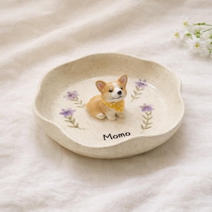 Może przedstawiać: Ceramiczna miska dla zwierząt w kolorze beżowym, z figurką corgi w ż&oacute;łtej apaszce w kropki. Miska jest ozdobiona fioletowymi akcentami kwiatowymi i napisem "Momo".