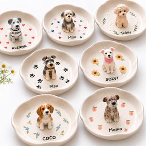 May include: A collection of seven round, off-white ceramic trinket dishes, each featuring a small, hand-painted dog figurine in the center. The dishes are personalized with names including Alenna, Milo, Teddy, Max, Solvi, and Momo, alongside motifs like hearts, flowers, paw prints, and peaches.