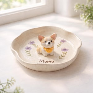 Może przedstawiać: Beżowy ceramiczny talerz z falistym brzegiem, z małą figurką corgi w ż&oacute;łtej chuście. Talerz jest ozdobiony fioletowymi kwiatowymi akcentami i napisem "Momo".