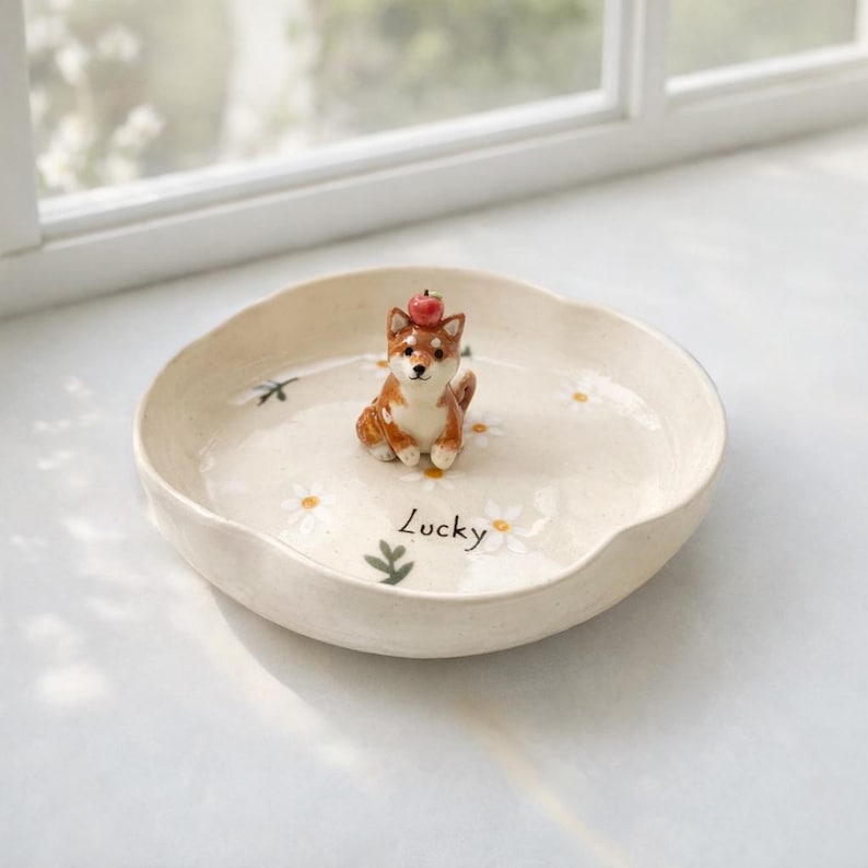 May include: A cream ceramic dish with a scalloped edge, decorated with daisies and a small brown and white Shiba Inu figurine wearing a red apple. The word "Lucky" is written on the dish.