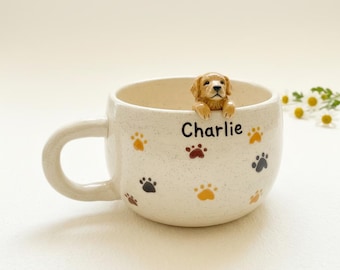 May include: Cream-colored ceramic mug with a handle, featuring a 3D golden retriever figurine peering over the rim. The mug is decorated with paw prints in various colors and the name "Charlie".