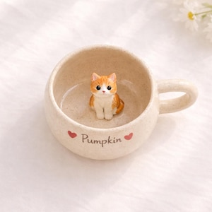 Puede incluir: Una taza de cerámica beige con una pequeña figura de gato naranja y blanco en su interior. La taza tiene un asa y la palabra "Pumpkin" impresa en la parte delantera, flanqueada por dos corazones rojos. La taza está sobre una superficie blanca.