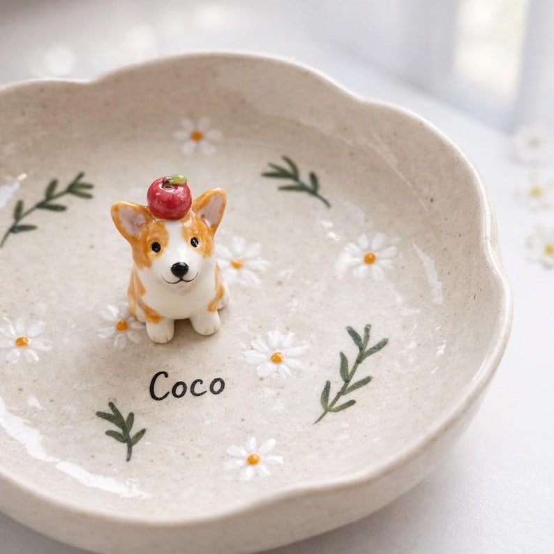 May include: A ceramic dish with a scalloped edge, decorated with white daisies and green sprigs. A small corgi figurine with an apple on its head sits in the centre. The name "Coco" is written on the dish.