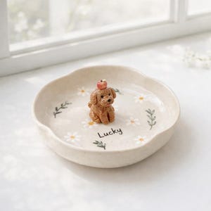 May include: A beige ceramic dish with a scalloped edge, decorated with white daisies and green leaves. A small brown dog figurine with an apple on its head sits in the center. The word "Lucky" is written on the dish.
