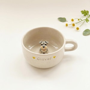 Puede incluir: Una taza de cerámica beige con una figura en miniatura de Schnauzer en su interior. La taza tiene un asa y la palabra "Clover" está escrita en el lateral con acentos de corazón amarillo. Un artículo decorativo encantador.