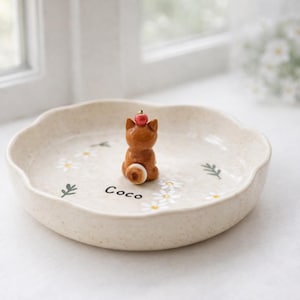 May include: A beige ceramic plate with a scalloped edge, decorated with white daisies and green leaves. A small brown cat figurine with a red apple on its head sits in the centre. The name "Coco" is written on the plate.