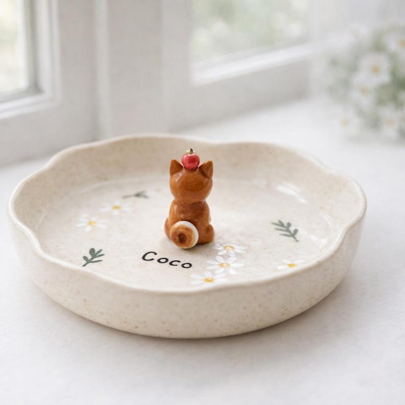 May include: A beige ceramic plate with a scalloped edge, decorated with white daisies and green leaves. A small brown cat figurine with a red apple on its head sits in the centre. The name "Coco" is written on the plate.