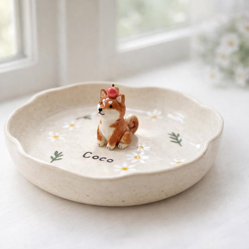 May include: A cream-coloured ceramic dish with a wavy edge, decorated with painted daisies and green sprigs. A small, brown and white Shiba Inu figurine with a red apple on its head is in the centre. The word "Coco" is written on the dish.