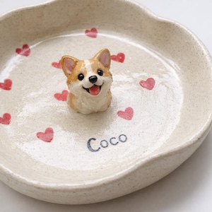 May include: A beige ceramic pet bowl featuring a corgi dog figurine and red heart decorations. The dog is tan and white, with a smiling face. The name "Coco" is written on the bowl.