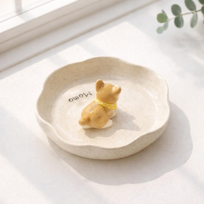 Może przedstawiać: Beżowy ceramiczny talerz z falistą krawędzią, z małą, brązową figurką psa z ż&oacute;łtym kołnierzem w kropki. Na talerzu wydrukowano słowo "Momo". Talerz znajduje się na białej powierzchni.