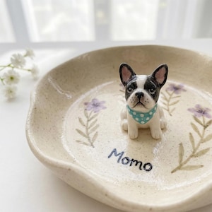 Może przedstawiać: Beżowa ceramiczna miska z czarno-białą figurką buldoga francuskiego w niebieskiej apaszce w kropki. Miska jest ozdobiona fioletowymi wzorami kwiatowymi i napisem "Momo".