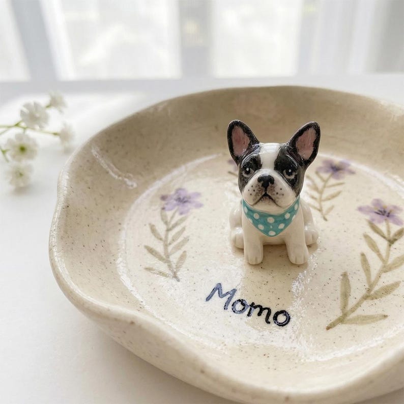 Może przedstawiać: Beżowa ceramiczna miska z czarno-białą figurką buldoga francuskiego w niebieskiej apaszce w kropki. Miska jest ozdobiona fioletowymi wzorami kwiatowymi i napisem "Momo".
