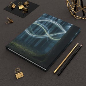 Peut inclure: Un journal à couverture rigide avec une scène de forêt et des motifs lumineux abstraits bleus et jaunes. Des fournitures de bureau dorées et noires, dont des trombones et des crayons, sont disposées sur une surface gris foncé.