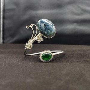 Puede incluir: Una pulsera plateada con dos piedras preciosas ovaladas. La piedra más grande es de color azul verdoso oscuro, y la más pequeña es de color verde vibrante. La pulsera tiene un elemento de diseño floral.