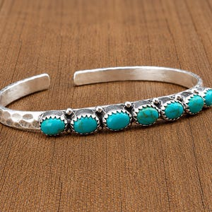 May include: A silver cuff bracelet featuring a hammered texture on the band and a row of six oval-shaped turquoise stones set in scalloped bezels. The open-ended design showcases a polished metallic finish with bright blue-green gemstone accents.