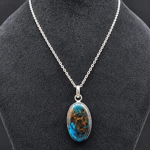 Peut inclure: Un collier à chaîne en argent avec un pendentif ovale en pierre polie. La pierre présente un mélange vibrant d'inclusions bleu profond et brun terreux, sertie dans un cadre en argent lisse relié à la fine chaîne à maillons en argent.