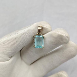 May include: A rectangular, light blue gemstone pendant with a gold-colored bail. The gemstone is set in a gold-colored frame. The pendant is held in a white glove.
