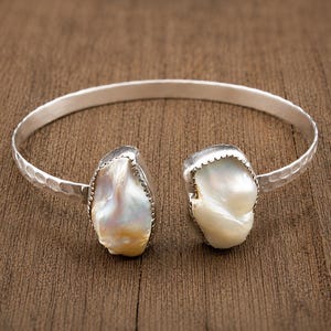 Puede incluir: Una pulsera de plata tipo brazalete con textura martillada en la banda y dos perlas barrocas blancas e iridiscentes de forma irregular, engastadas en biseles de plata dentados en cada extremo abierto, creando una pieza de joyería brillante.