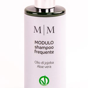 Op de afbeelding: Cilindrische shampoo fles met een zilveren dop en een donkergroene hals. Het etiket toont de letters "MM" boven de woorden "MODULO shampoo frequente". Daaronder staat "Olio di jojoba Aloe vera" en "MADE IN ITALY WITH LOVE".