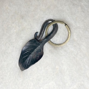May include: A handcrafted metal leaf keychain with a dark finish. The leaf design is detailed with etched veins and is attached to a brass-colored keyring. A unique accessory for keys or bags.