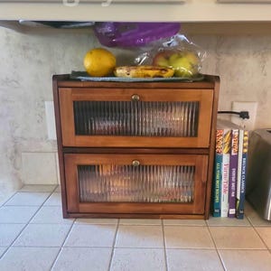 May include: A two-drawer wooden bread box with glass paneled doors. The bread box is sitting on a tiled countertop. The text "VintagByDeni" is visible in the image. A toaster and some books are also visible.