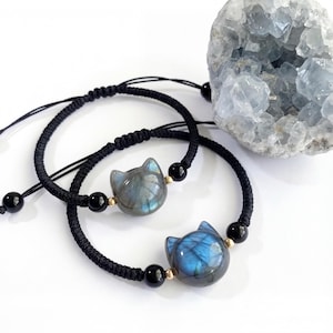 May include: Two black cord bracelets, each featuring a cat-shaped labradorite stone with blue and gray hues. Accented with black and gold beads. The bracelets are displayed on a white surface.
