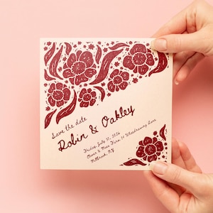 May include: A square cream-colored save the date card featuring dark red floral illustrations in the corners. The text reads: Save the date, Robin & Oakley, Friday, July 31, 2026, Clover & Moss Farm 14 Wheedrawing Lane, Millbrook, NY.
