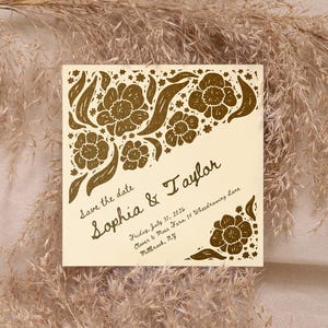 May include: A square cream-colored save the date card featuring gold floral illustrations in the corners. The text reads: Save the date, Sophia & Taylor, Friday, July 31, 2026, Clover & Moss Farm 14 Wheedrawing Lane, Millbrook, NY.