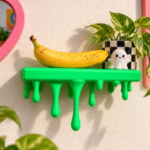 May include: A bright green wall shelf with a decorative dripping paint design. A yellow banana, a small white ghost figurine with a smiling face, and a black and white checkered planter containing a green leafy plant sit on the shelf surface.