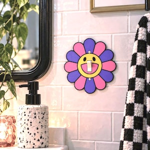 May include: A decorative light switch cover shaped like a flower with alternating purple and pink petals. The center of the flower features a yellow face with a wide, smiling mouth and black outlines. The cover is mounted over a white toggle switch.