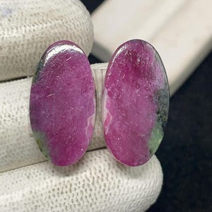 May include: Two oval-shaped gemstones with a polished surface. The stones are primarily a deep magenta color, with areas of green and black. The gemstones are displayed against a neutral background.