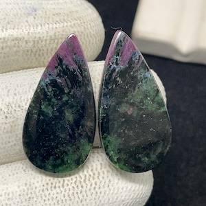 Könnte beinhalten: Zwei tropfenförmige Edelsteine. Die polierten Steine weisen eine Mischung aus Farben auf, darunter tiefes Grün, Schwarz und Flecken von leuchtendem Rosa und Lila. Die Edelsteine ​​sind vor einem weißen Hintergrund platziert.