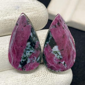 May include: Two teardrop-shaped gemstones with a vibrant pink hue, interspersed with dark green and white patterns. The polished stones have a smooth surface and are displayed against a neutral background, showcasing their natural beauty.