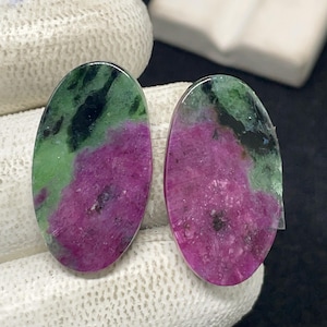 May include: Two oval-shaped gemstones with a polished surface. The stones feature a vibrant mix of colors, including deep magenta, forest green, and black. The gemstones are likely intended for jewelry making or collection.