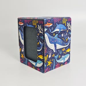 May include: A rectangular box with a dark blue background featuring a colorful pattern of whales, small fish, and coral reefs in shades of light blue, yellow, and pink. The front of the box has a centered, vertical rectangular cutout opening.