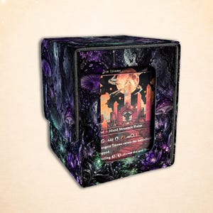 Op de afbeelding: Een vierkante deckbox met een donkerpaarse en zwarte bosillustratie met lichtgevende paddenstoelen. Een rechthoekig venster aan de voorzijde toont een ruilkaart met een landschap, een grote maan, kristallen en gedrukte tekst.