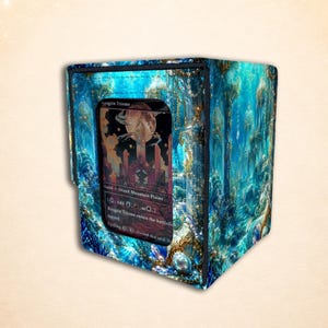May include: A rectangular deck box featuring a vibrant blue and teal underwater forest scene with gold accents. A clear window on the front displays a Raugrin Triome trading card. The box has black trim along the edges and a structured design.