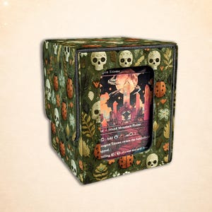 Op de afbeelding: Een groene kaartenopbergdoos met een herhalend patroon van witte schedels, lieveheersbeestjes en bladeren. De doos bevat een kaart met een fantasielandschapsillustratie. De tekst "grin Triome" is zichtbaar op de kaart.