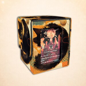 May include: A square card storage box featuring a black cat illustration on an orange and cream checkered background. The front has a rectangular cutout displaying a trading card with a landscape illustration and the text Savai Triome.