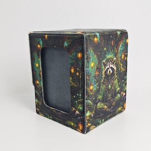 May include: A rectangular deck box featuring a textured surface with a printed illustration of a raccoon sitting in a forest setting. The box has a dark, solid-colored rectangular cutout on the front panel and a hinged lid design.