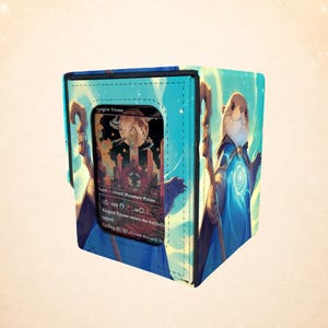 Op de afbeelding: Blauwe kaartenopbergdoos met een cartoon otter tovenaar met een staf. De doos heeft een zichtbare kaart met de tekst "Angrin Triome". De doos is versierd met een sterrenhemel en een volle maan.