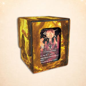 Puede incluir: Una caja para barajas rectangular con acabado amarillo dorado que presenta la silueta de una criatura sosteniendo un paraguas en el lateral. El panel frontal tiene una ventana transparente que muestra una carta con el texto Savai Triome.