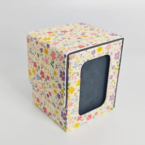 Op de afbeelding: Een vierkante opbergdoos met een crèmekleurige buitenkant en een dicht, veelkleurig bloemenpatroon in tinten roze, geel, blauw en paars. Het voorpaneel bevat een rechthoekige uitsparing die een donkergekleurde binnenkant laat zien.