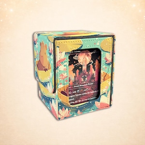 Puede incluir: Una caja de almacenamiento de tarjetas cuadrada con un colorido diseño floral y capibara. La caja tiene una tarjeta visible con el texto "grin Triome" y "Island Mountain Plains". La caja está hecha de tela y tiene un cierre magnético.