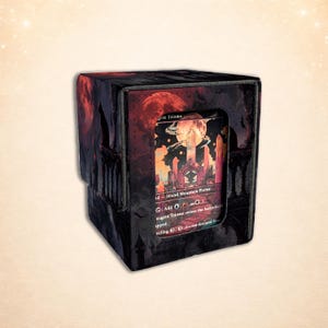 May include: A black card storage box with a red and black fantasy landscape design. The box has a card visible with the text "Grin Triome" and a landscape illustration. The box is designed to hold playing cards.