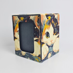 May include: A square-shaped desk organizer or pen holder featuring a colorful graphic print of a calico cat with large eyes against a dark blue space background with stars and planets. The item has a black interior and a rectangular cutout on the front.